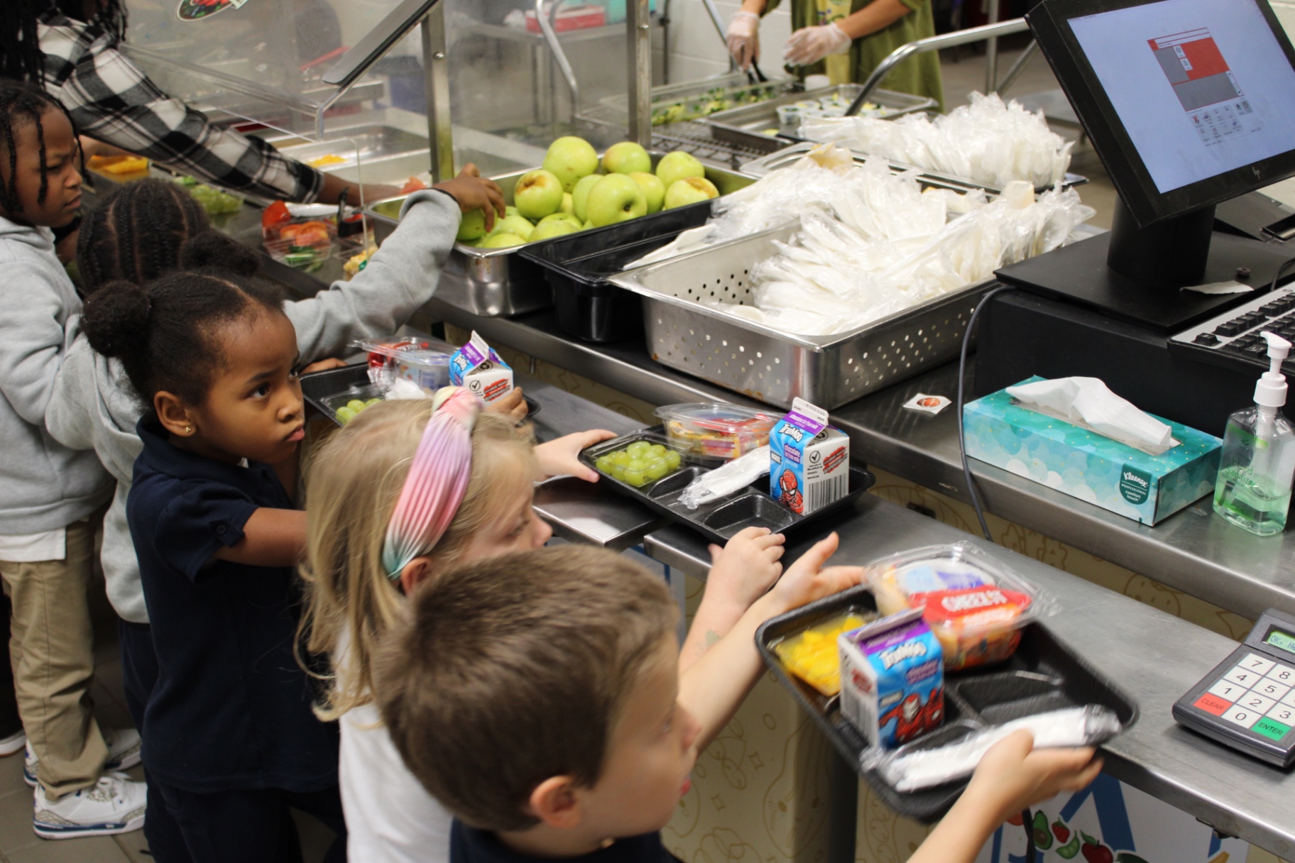 APS elementary students selecting fresh fruit and carrying complete school meals through a cafeteria service line